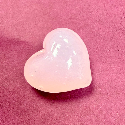 A polished, heart-shaped translucent stone, likely rose quartz, resting on a textured pink surface.