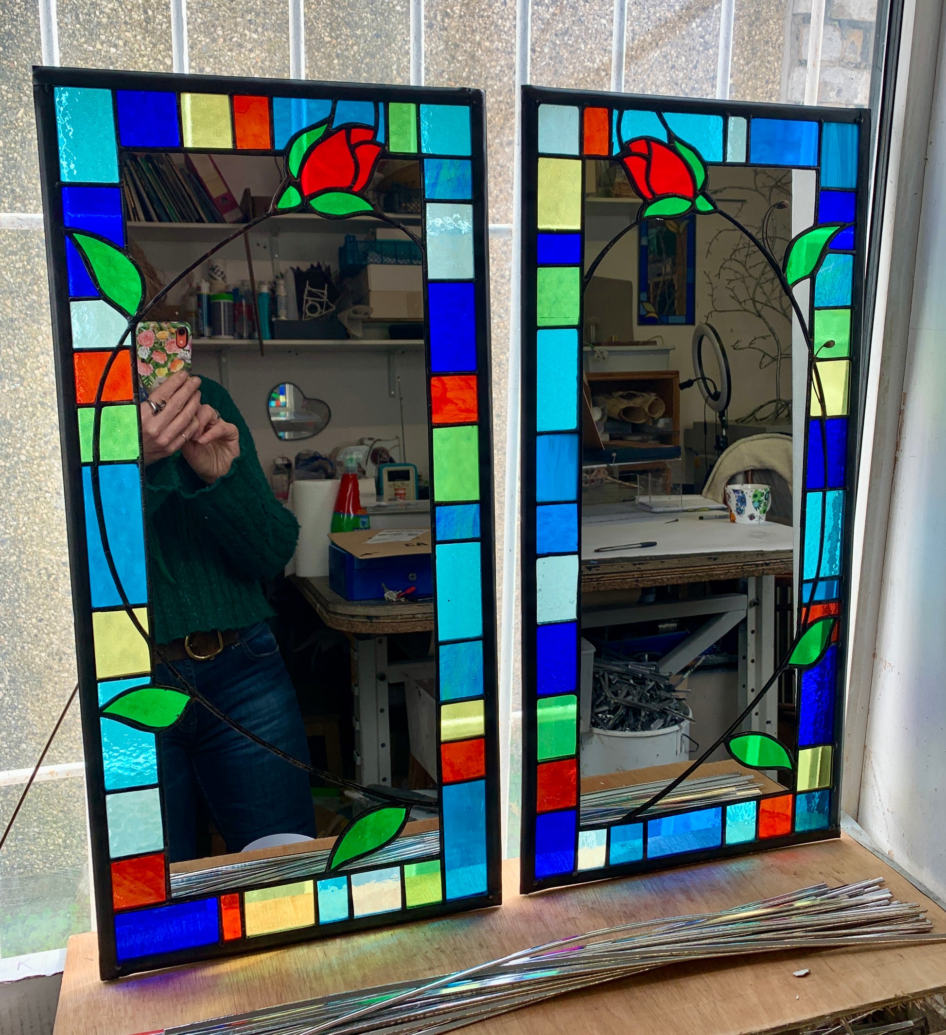 Tall slim stained glass mirror with random colours in various sizes in the border. Red rose bud detail with a long stem cut into the mirror glass and border. Copperwire attached to the back for hanging.