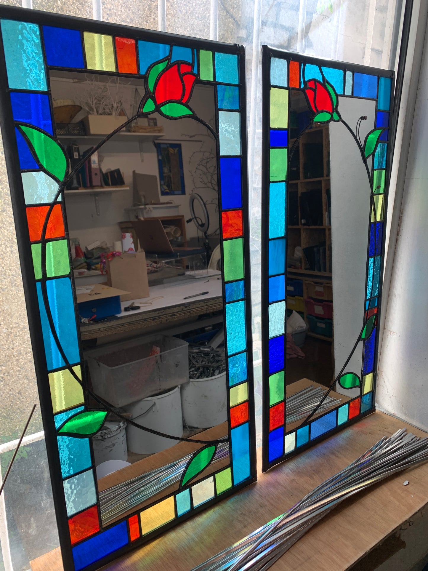 Tall slim stained glass mirror with random colours in various sizes in the border. Red rose bud detail with a long stem cut into the mirror glass and border. Copperwire attached to the back for hanging.