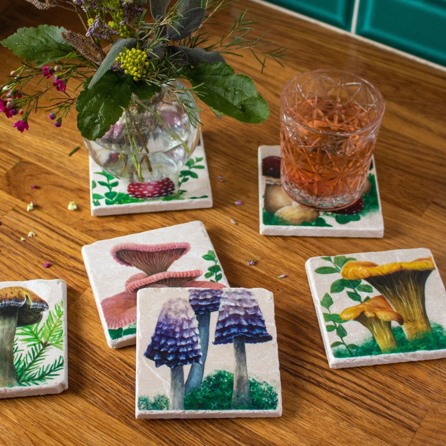 An assortment of six stone coasters with different hand painted woodland mushroom designs with an inkcap mushroom design in front. On the upper right hand side a drink rests on top of one of the coasters. On the top left there is a round vase of flowers on top of a coaster.