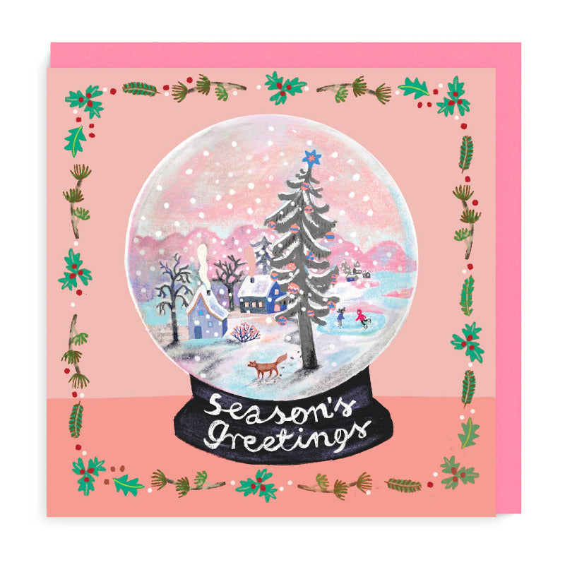 Christmas card in a folk art style with a snow covered village in a snow globe and a pink background with holly and misletow