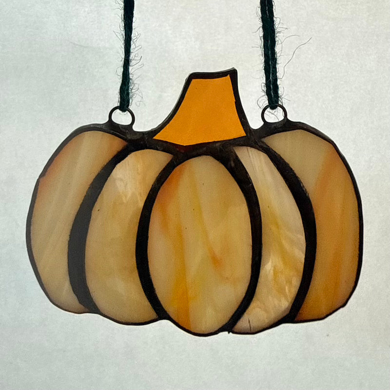 A stained glass pumpkin ornament with a textured orange and yellow glass body, dark black metal outlines, and a black hanging cord, shown against a light gray background.