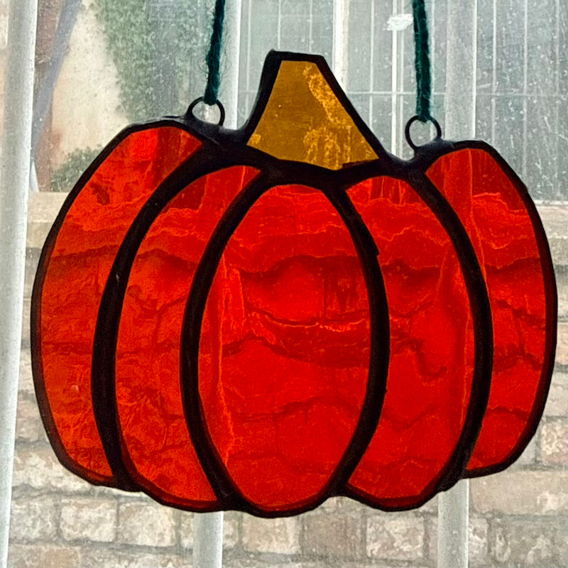 A stained glass pumpkin ornament with a textured red body, a gold stem, and black metal outlines, hanging in front of a window.