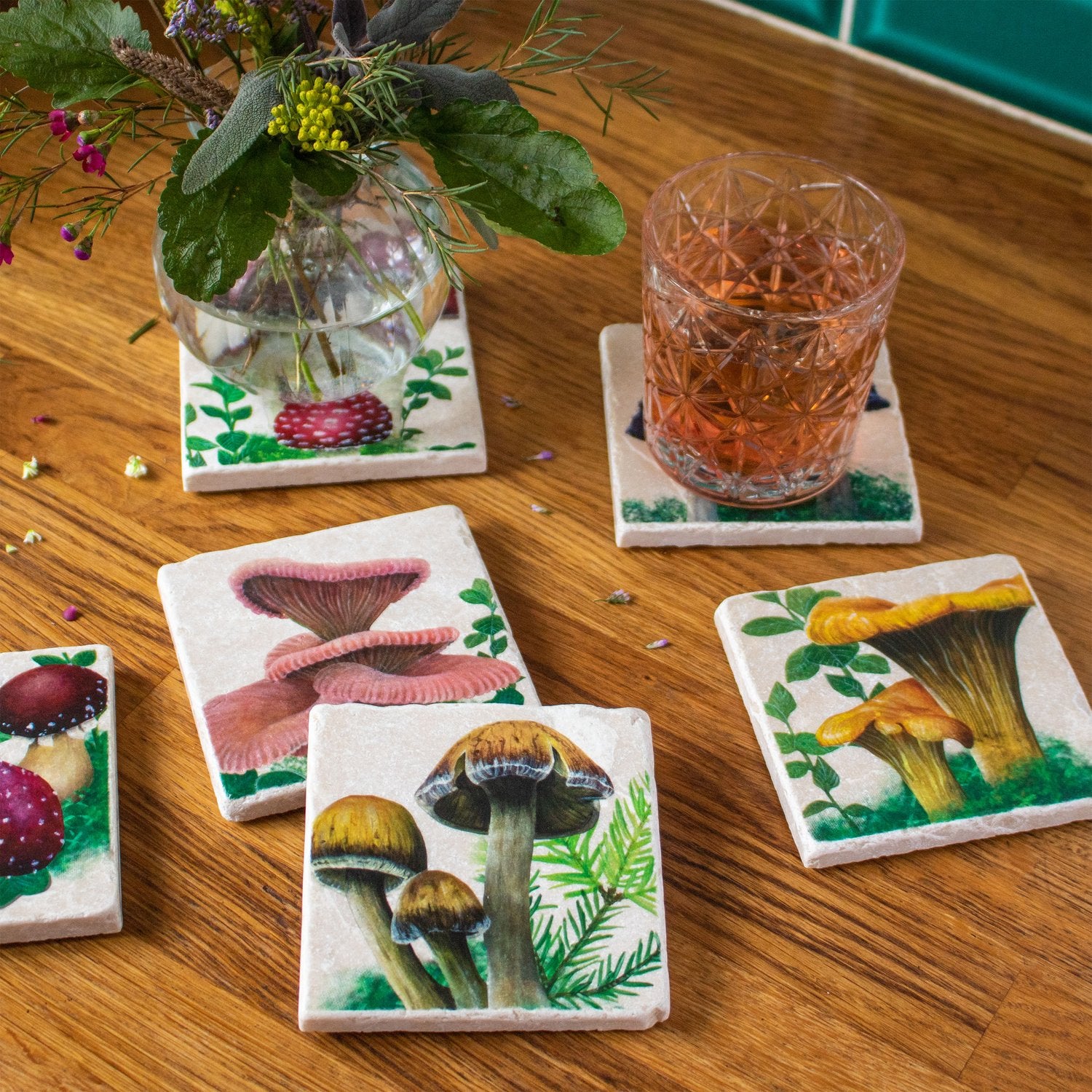An assortment of six stone coasters with different hand painted woodland mushroom designs with a parasol mushroom design in front. On the upper right hand side a drink rests on top of one of the coasters. On the top left there is a round vase of flowers on top of a coaster.
