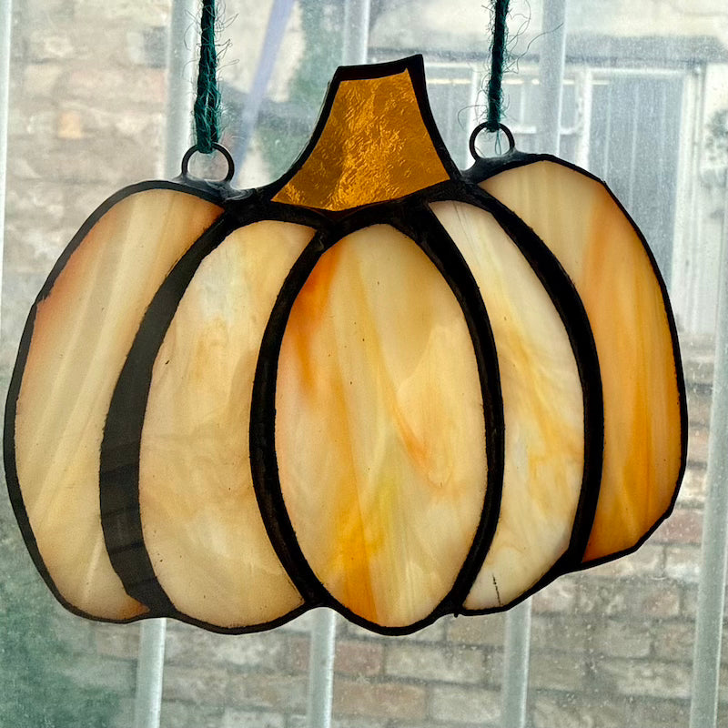A stained glass pumpkin hanging ornament with a textured amber glass stem and marbled cream, orange, and yellow glass panels divided by black solder lines, suspended by dark green cords in front of a window.
