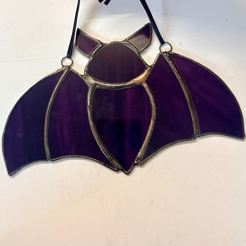 A stained glass bat ornament with deep purple glass panels and silver metal leading, suspended by a black string against a plain off-white background.