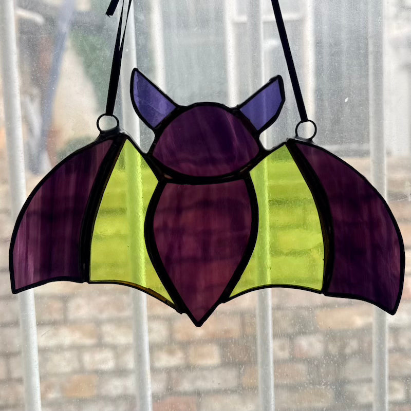 A stained glass bat suncatcher with purple wings and body, and bright yellow-green wings, hanging on a window.