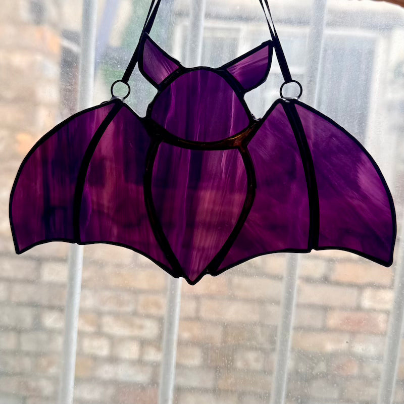A purple stained glass suncatcher shaped like a bat, suspended by black cords on a window with a blurred brick wall background.