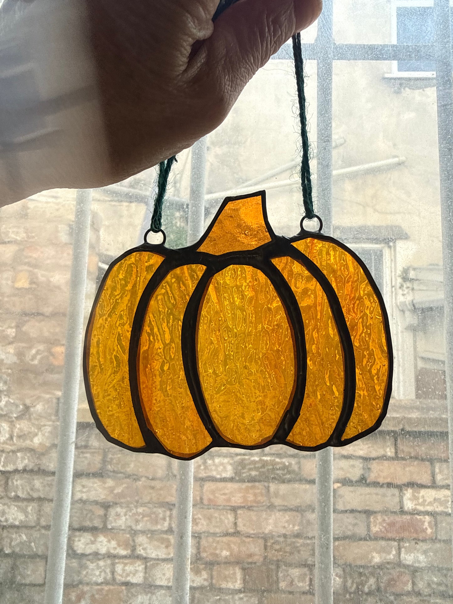 A hand holds a stained-glass pumpkin ornament with amber and orange panels outlined in black, suspended in front of a window with a brick wall visible outside.