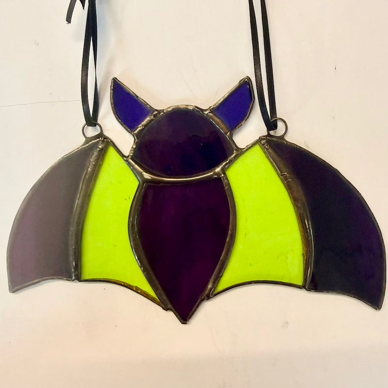 A stained glass bat ornament with purple, neon yellow, and dark gray glass panels, outlined in gold-toned metal and suspended by black hanging loops against a plain off-white background.