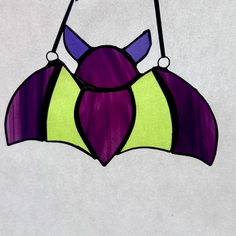 A bat-shaped hanging ornament or wall hanging with a stained-glass look, featuring purple wings and head, lime green body panels, and black hanging straps on a light gray background.