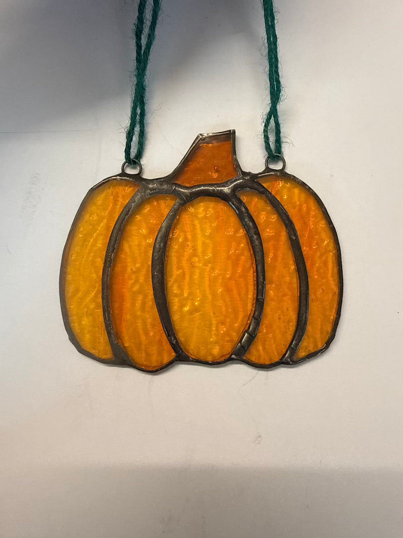 A stained glass pumpkin ornament with textured orange and yellow glass panels, dark metal outlines, and a dark green hanging string, shown against a plain off-white background.