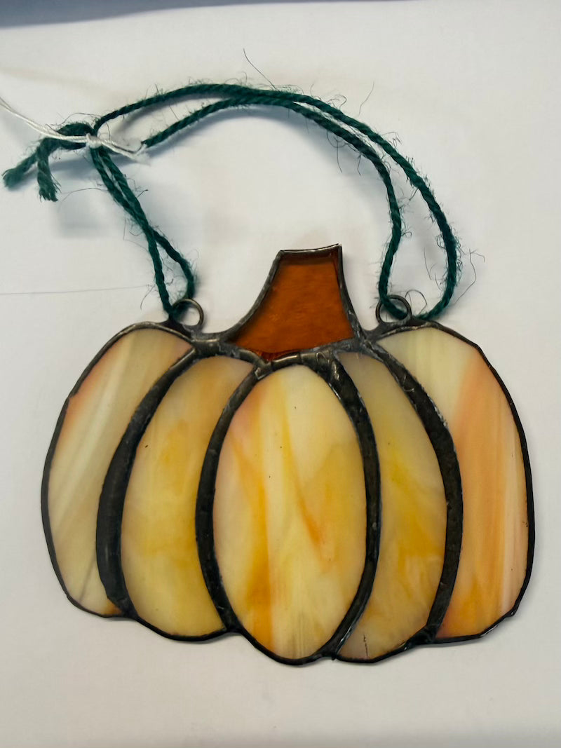 A stained glass pumpkin ornament with five vertical panes in cream, yellow, and orange tones, outlined in black with a dark green hanging string, shown on a plain white background.