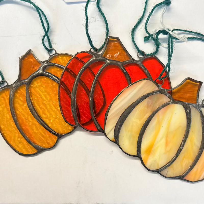 A set of five stained glass pumpkin ornaments in a warm autumn gradient, hanging on a white surface. The pumpkins transition from yellow-orange on the left, through red, to cream-white on the right, each with a dark green hanging string.