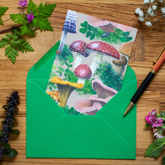 This bold Woodland Shrooms greeting card features a dense design of various mushrooms and fungi, surrounded by patches of moss. It has been printed on a sandy beige background. A bright red toadstool (or Fly Agaric) is sitting right in the middle of the card. The red Fly Agaric is surrounded by Chanterelles, Pink Oyster mushrooms, Inkcap mushrooms and an occasional snail or slug crawling through here and there.