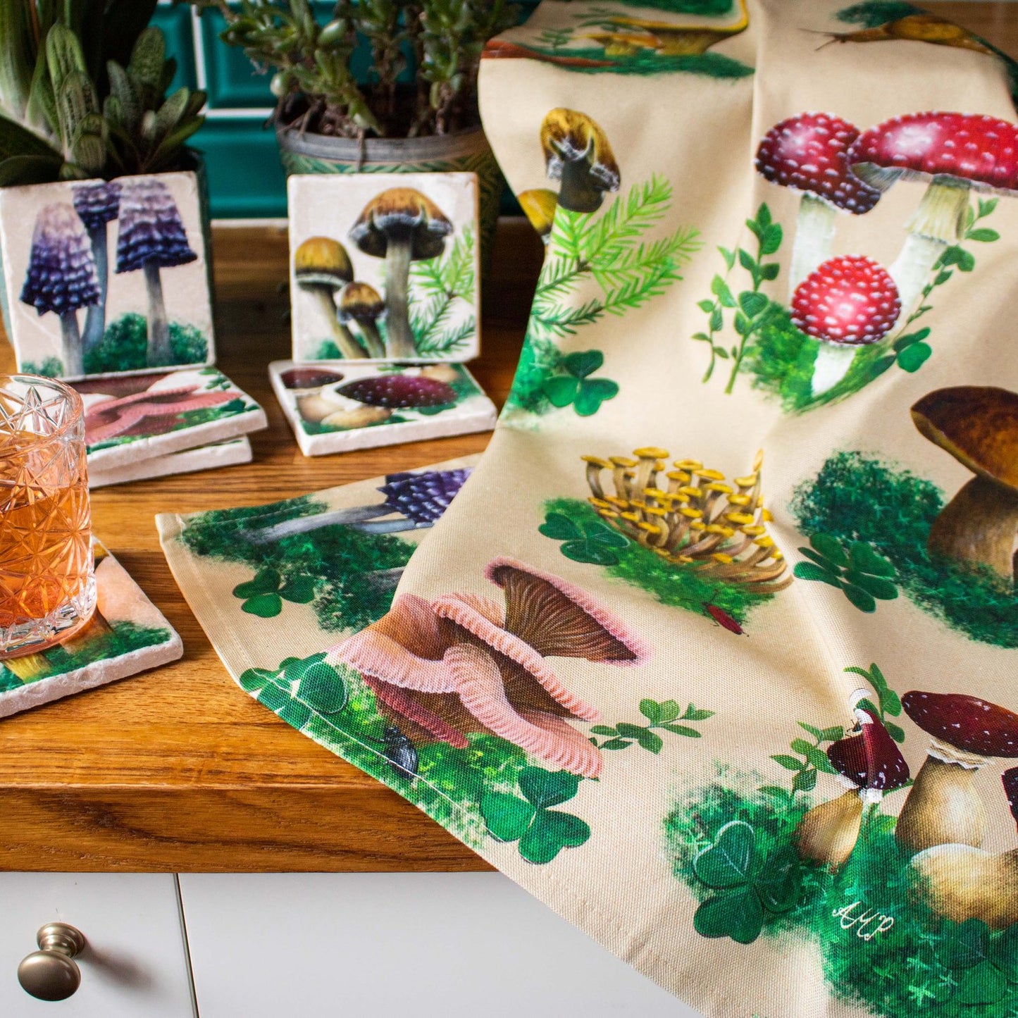A close up view of a tea towel with a print depicting various woodland mushrooms resting against a counter next to woodland mushroom themed stone coasters.