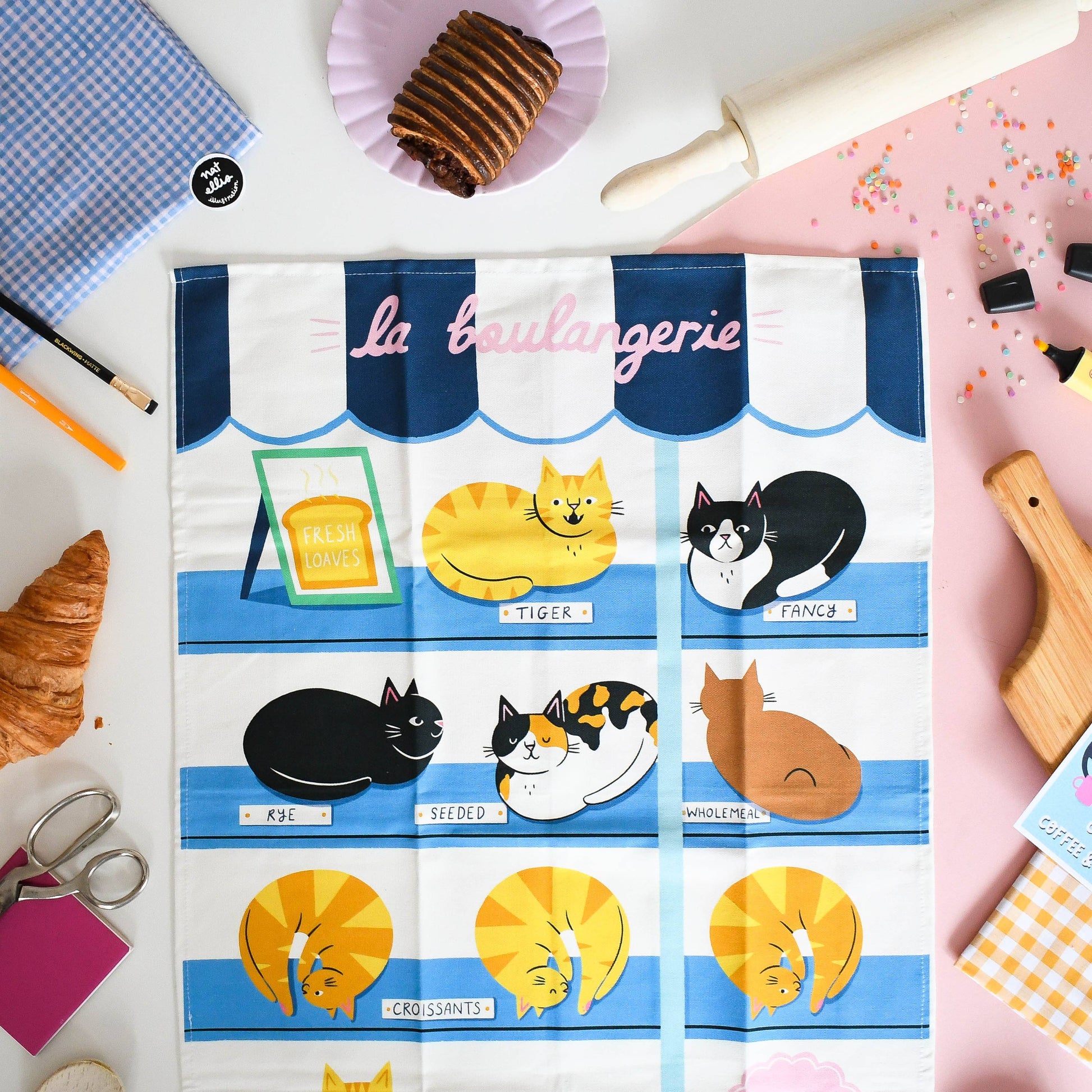 Tea towel with cat illustrations and 'la boulangerie' text on a white background with baking items.