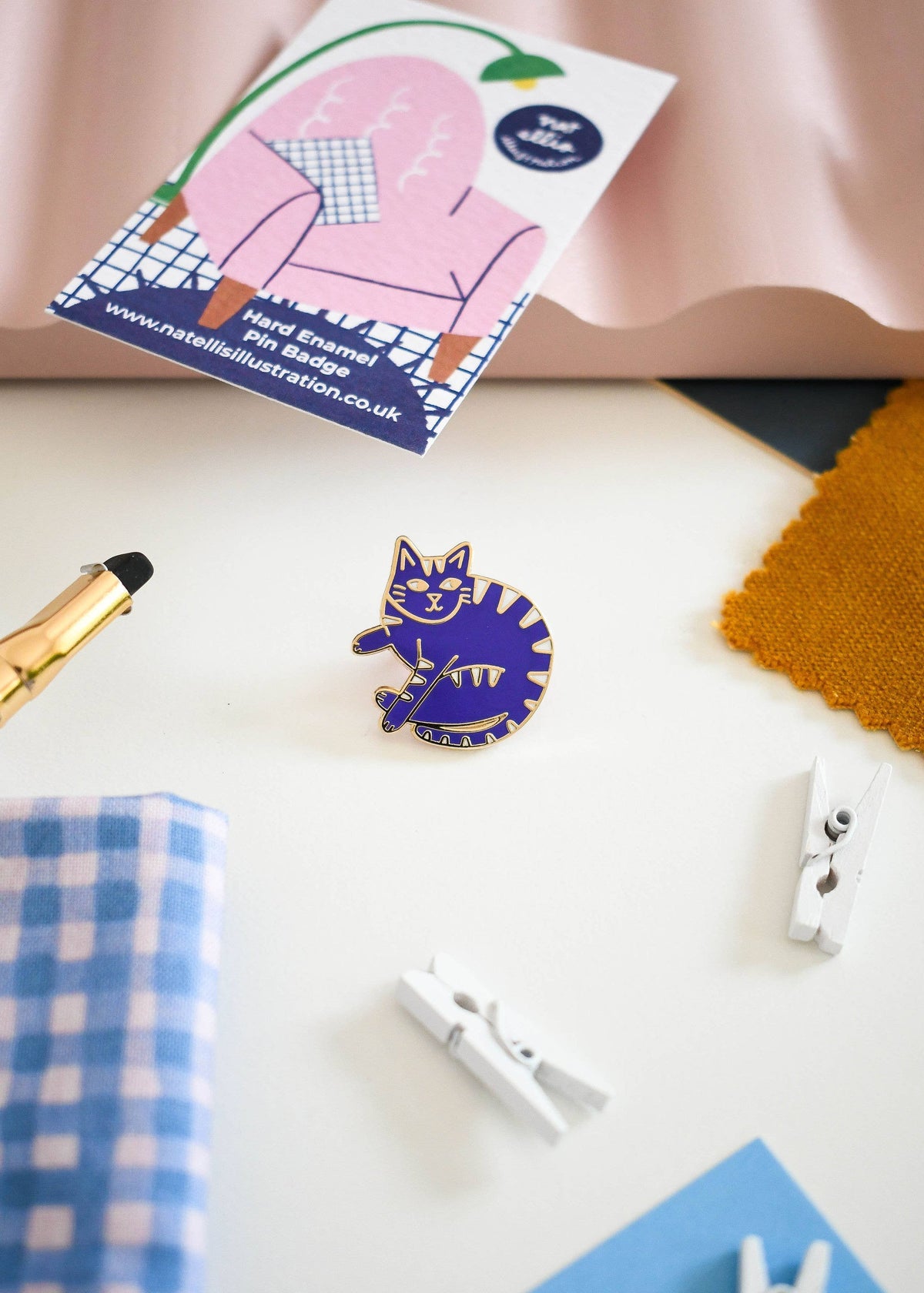 A small enamel pin shaped like a curled-up cat in blue and yellow stripes, displayed on a white surface with a branded card, fabric swatches, and small white clothespins around it.