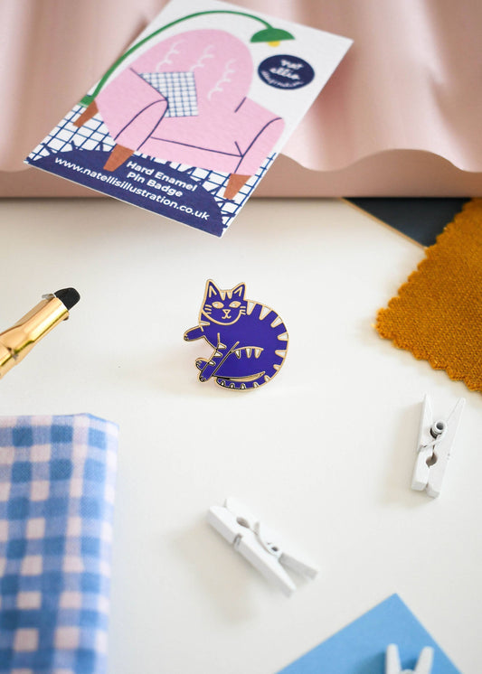 A small enamel pin shaped like a curled-up cat in blue and yellow stripes, displayed on a white surface with a branded card, fabric swatches, and small white clothespins around it.