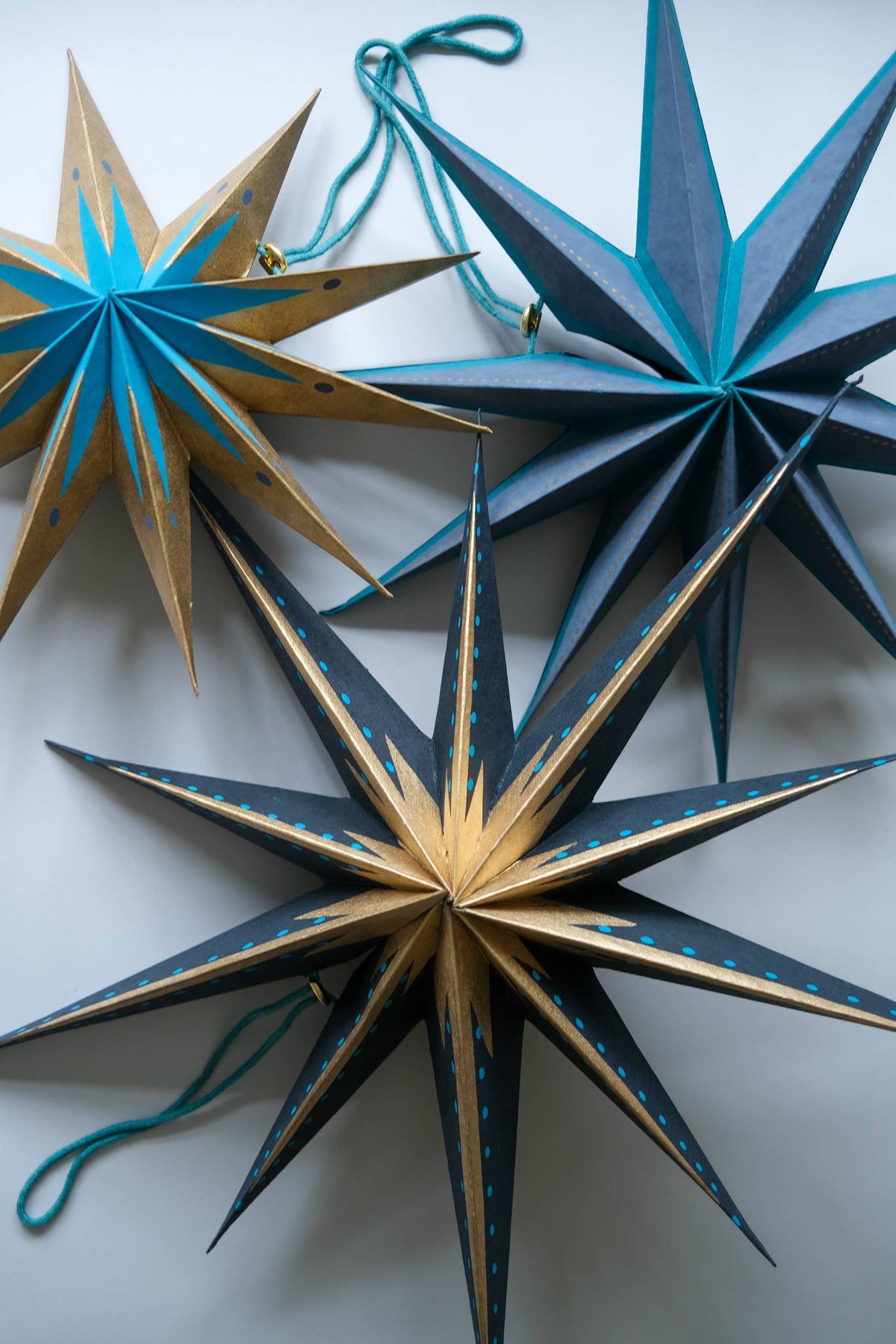 Three hand-painted wooden star ornaments hanging against a light gray background. The ornaments feature geometric star designs with alternating dark gray and gold panels, decorated with hand-painted blue stars and dots, and each has a teal hanging string.