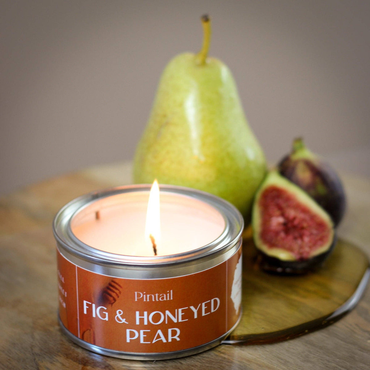 Candle in a 'Fig & Honeyed Pear' tin with a pear and figs in the background on a wooden surface.
