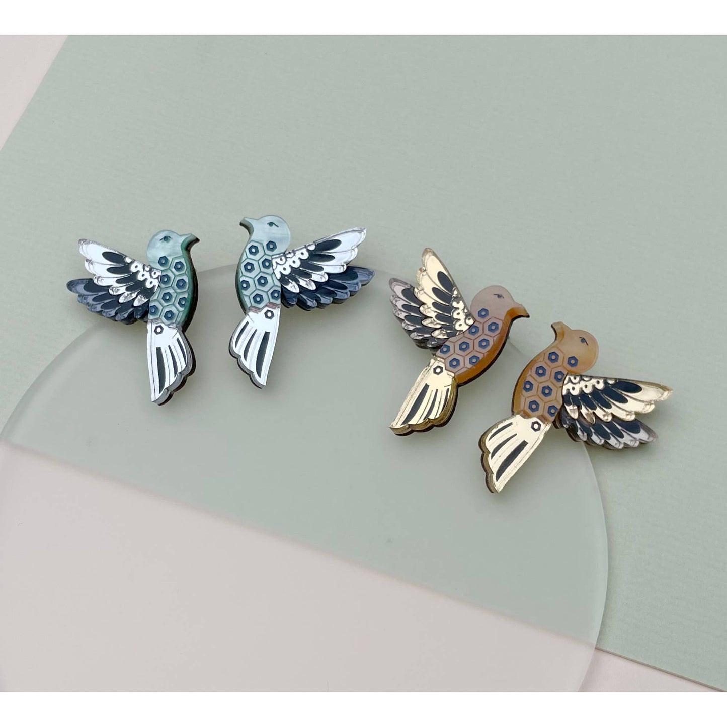 Four bird-shaped enamel pins arranged on a pale green and white surface. Two pins are light blue and white, and two are tan and cream with dark blue circular patterns and black outlines.