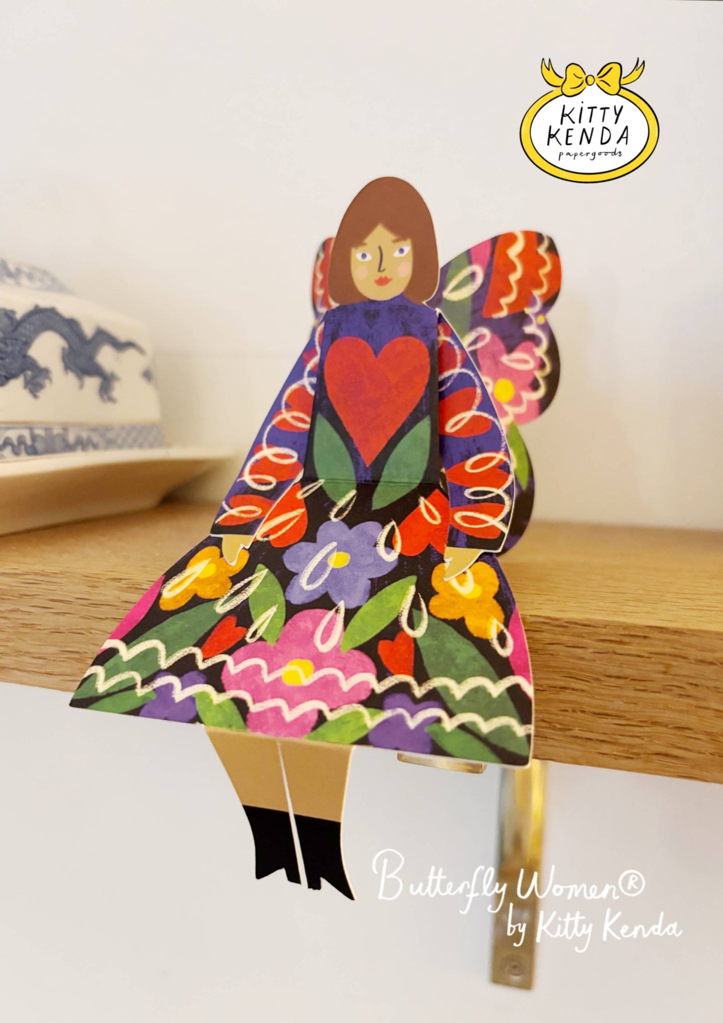Colorful butterfly-shaped figurine with a heart design on a wooden surface, branded 'Kitty Kenda'.