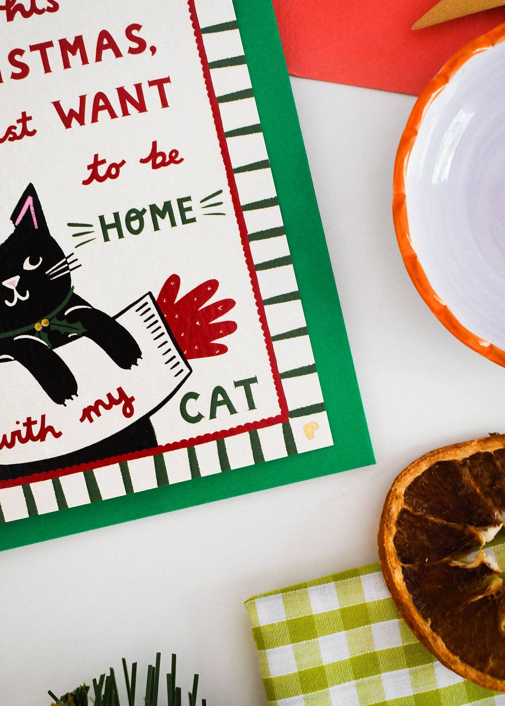 A festive Christmas-themed tea towel laid flat on a white surface, styled with a dried orange slice, a green-and-white checkered napkin, and a white bowl with an orange rim.
