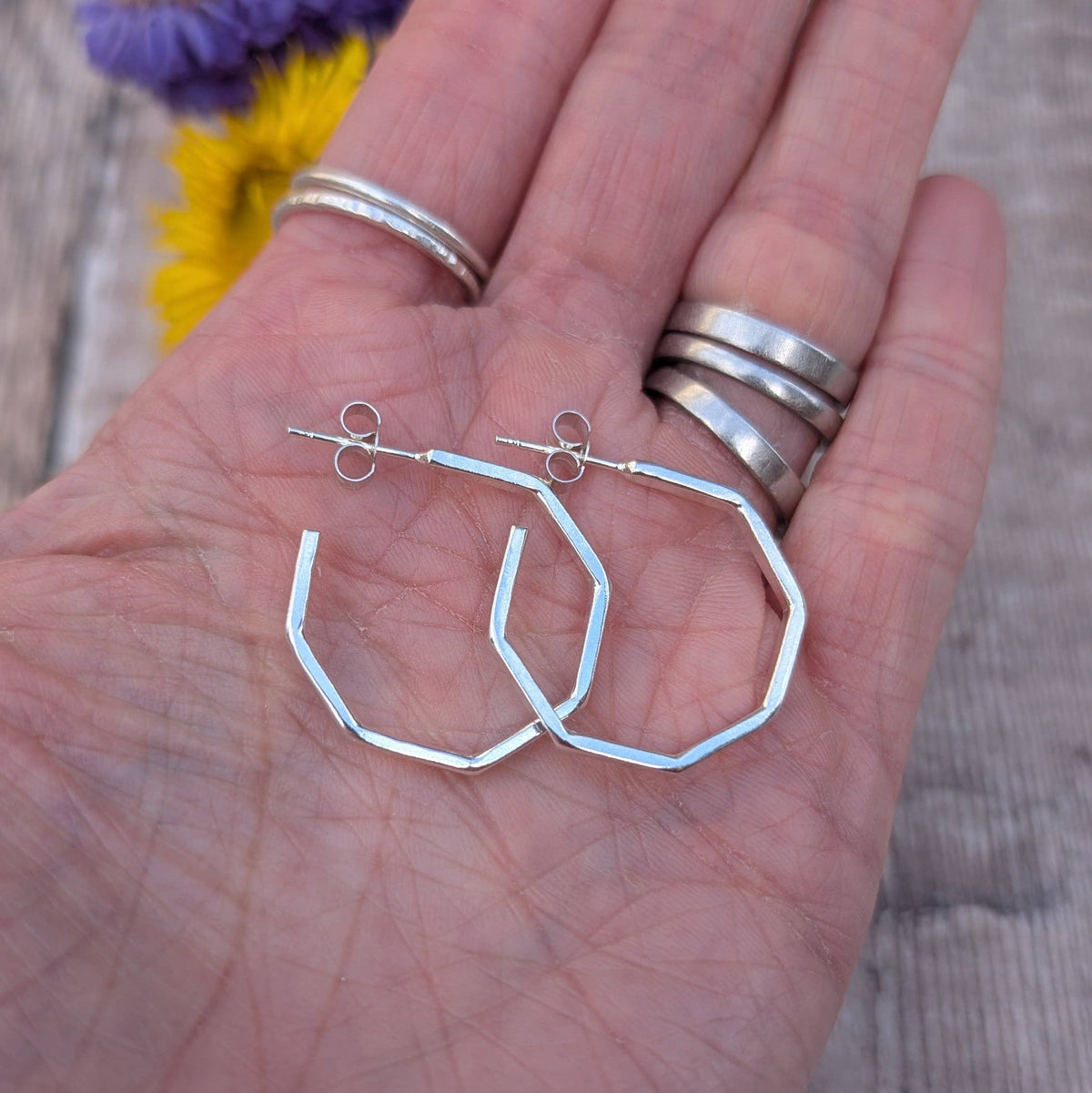 A pair of silver-toned geometric hoop earrings resting on an open hand, with several silver rings worn on the fingers and blurred flowers in the background.