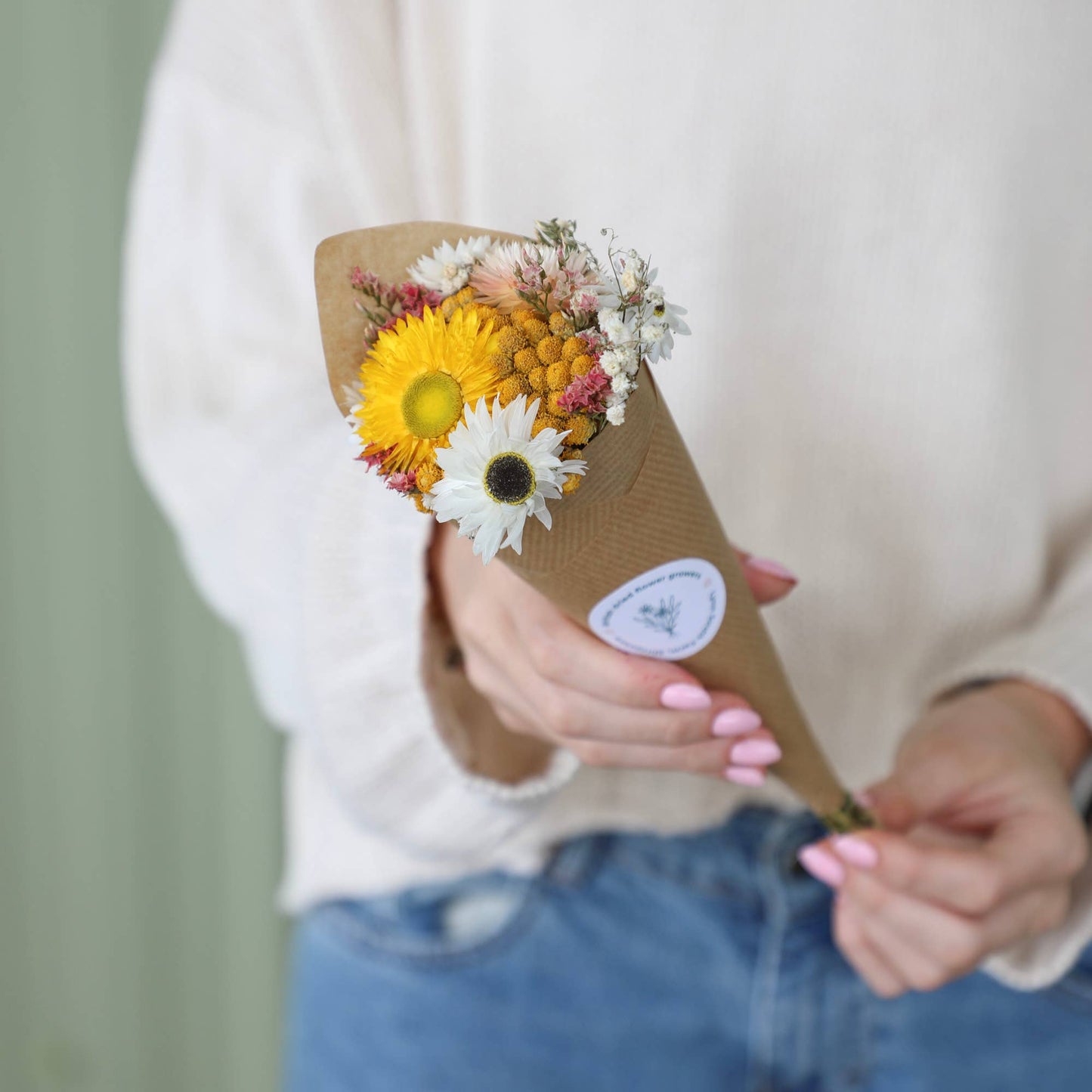 A person in a white sweater and blue jeans holds a small dried flower bouquet wrapped in brown kraft paper with a round white sticker. The bouquet includes yellow, white, and pink dried flowers.