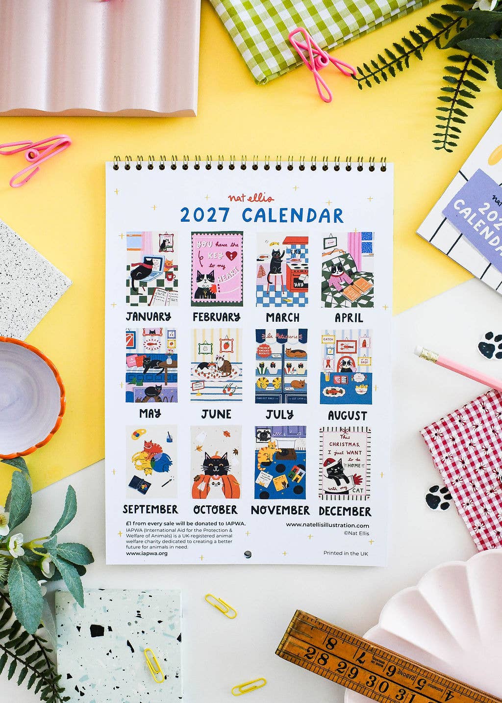 A spiral-bound 2027 calendar page titled 'rat ella 2027 CALENDAR' is laid flat on a bright yellow surface, surrounded by styled desk props including a pink bowl, green leaves, a wooden ruler, paper clips, and patterned notebooks.