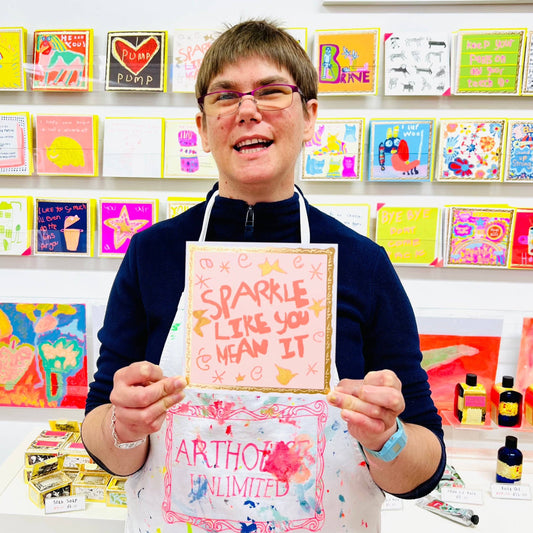 Person holding a colorful card with 'Sparkle like you mean it' text, wearing an apron in a card-making studio.