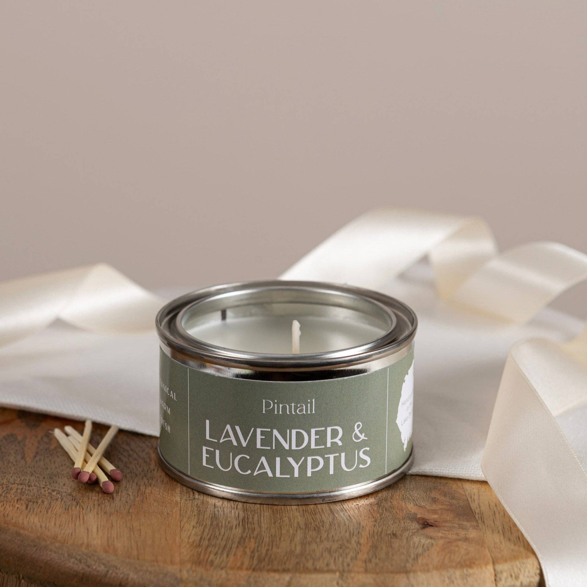 Candle in a tin labeled 'Lavender & Eucalyptus' by Pintail on a wooden surface with white fabric in the background.