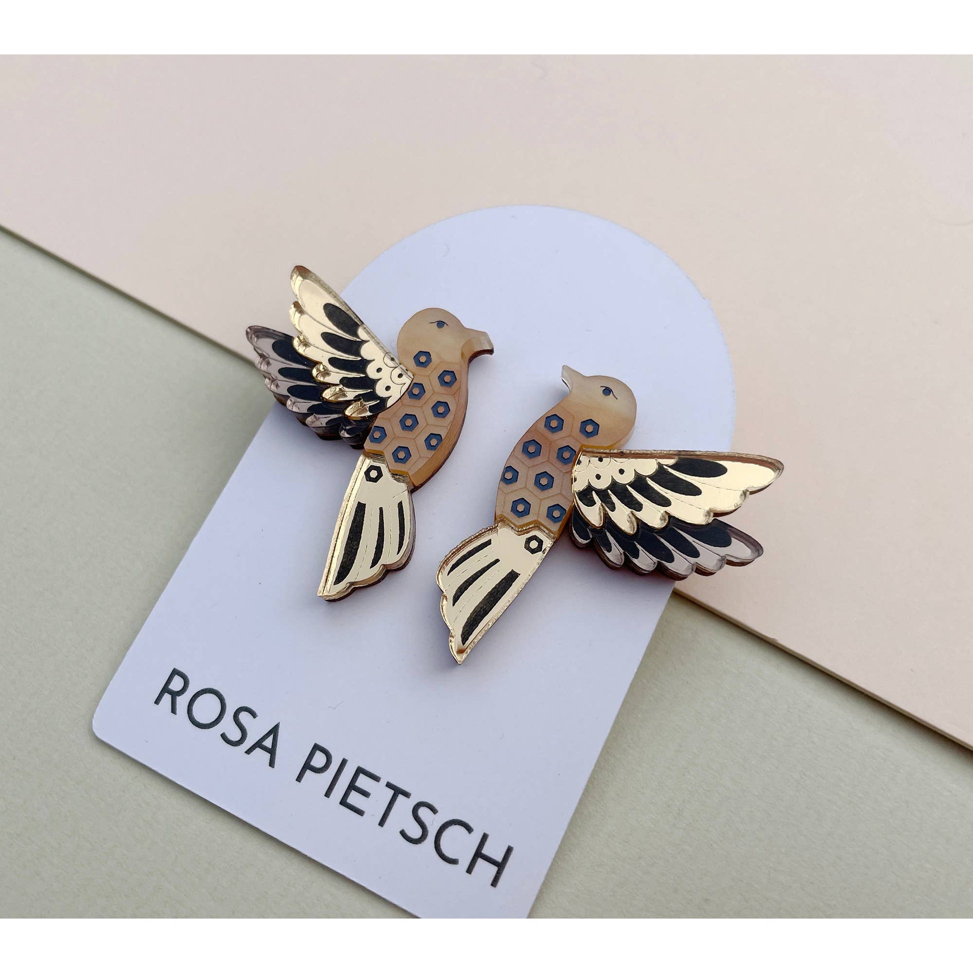 A pair of bird-shaped enamel pins displayed on a white card printed with the brand name 'ROSA PIETSCH'. The pins feature a gold-tone metal body with black and blue spotted details on the wings and body, set against a split beige and light green background.