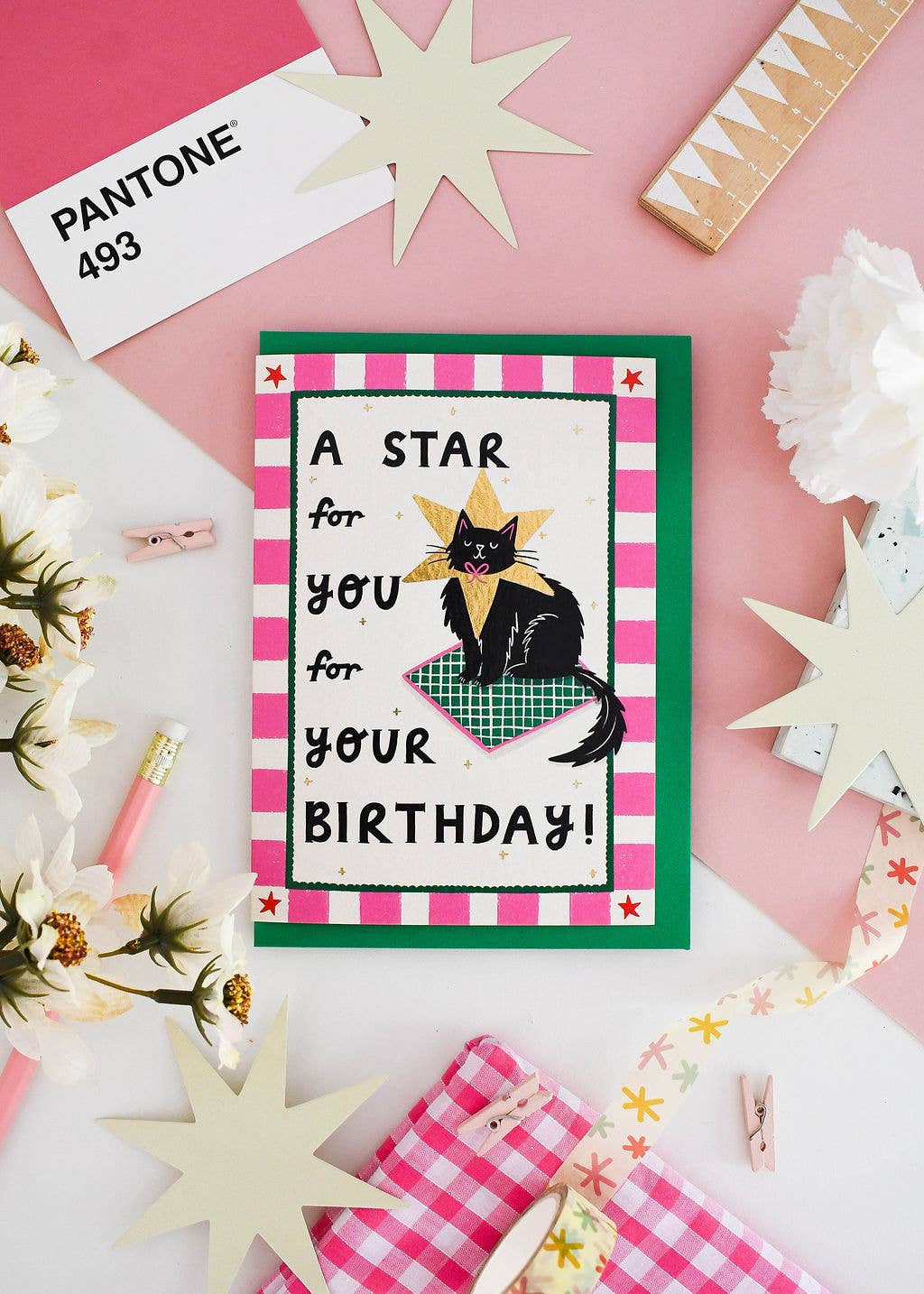 A handmade birthday card featuring a black cat wearing a gold star and a green plaid blanket, surrounded by pink and white checkered borders. The card is displayed in a styled flat-lay on a pink and white background with decorative paper stars, a Pantone card, and ribbon.