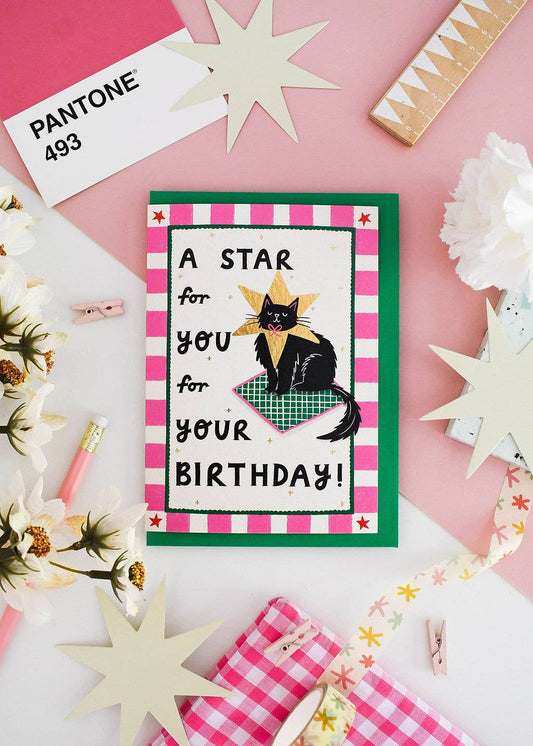 A handmade birthday card featuring a black cat wearing a gold star and a green plaid blanket, surrounded by pink and white checkered borders. The card is displayed in a styled flat-lay on a pink and white background with decorative paper stars, a Pantone card, and ribbon.