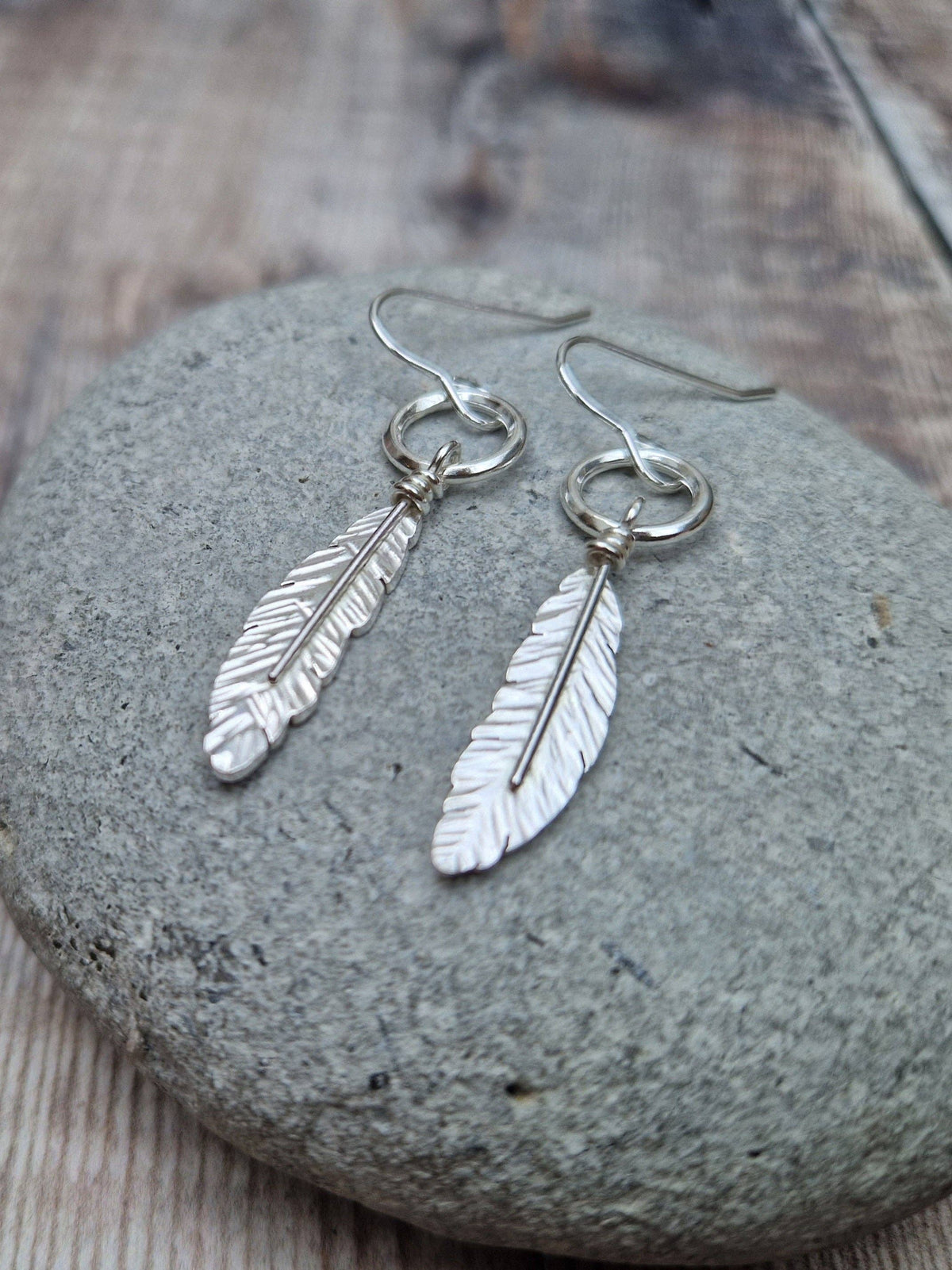 A pair of silver feather drop earrings displayed on a smooth gray stone over a rustic wooden surface.