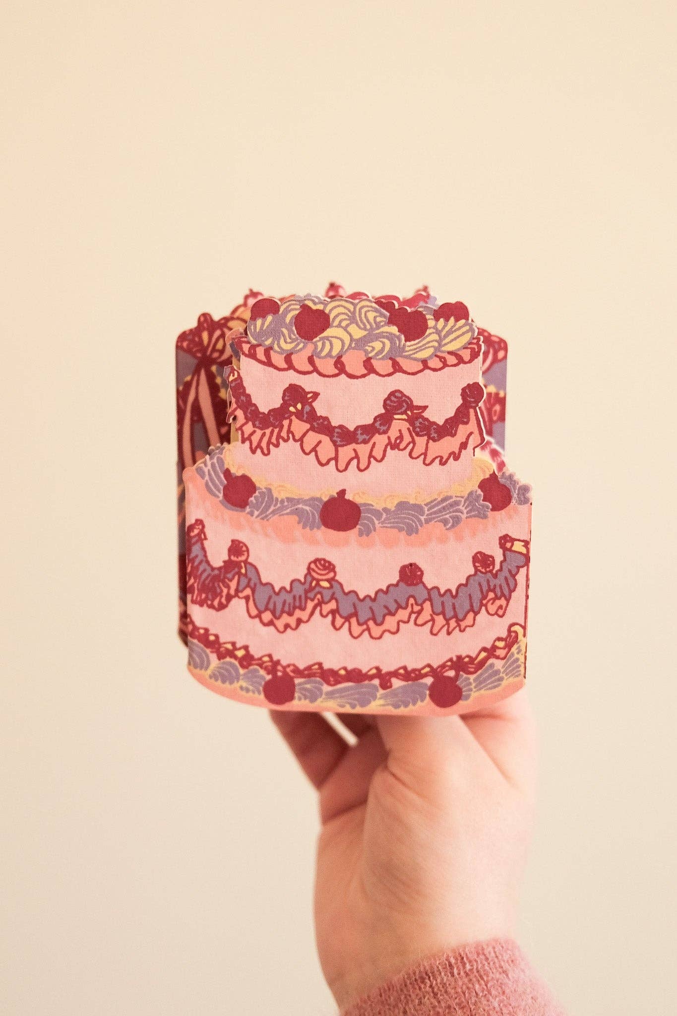 Hand holding a small, decorative cake-shaped object with intricate designs on a beige background