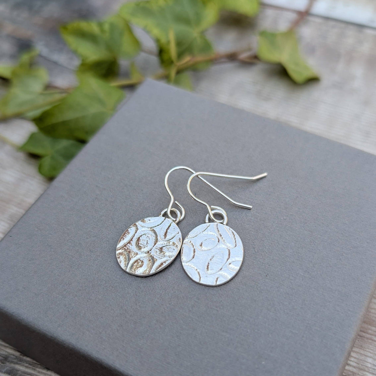 A pair of silver drop earrings with textured, organic circular cutouts, displayed on a dark gray square box with green leaves in the background.