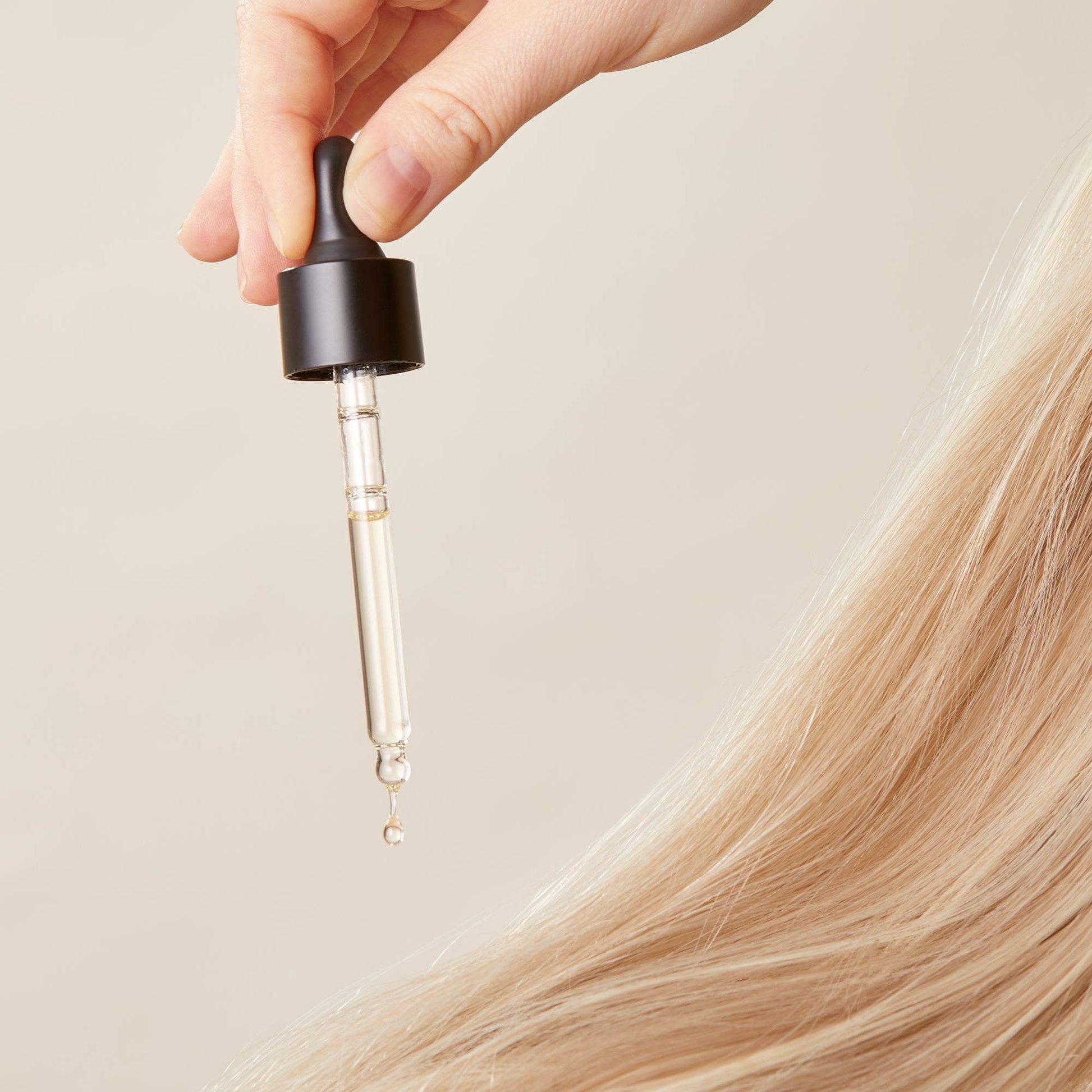 Hand holding a dropper with a hair oil over blonde hair on a neutral background