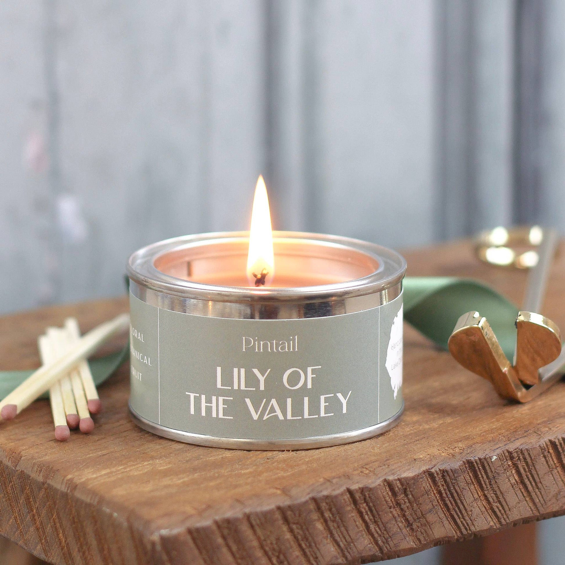 Candle labeled 'Lily of the Valley' by Pintail on a wooden surface with matches and a green container.