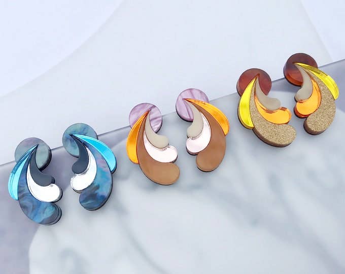 Four pairs of abstract layered stud earrings arranged on a light marble surface. The earrings feature curved, petal-like layers in different color combinations: one pair is blue, white, and gray; two pairs are orange, beige, and brown; and two pairs are yellow, orange, brown, and white.