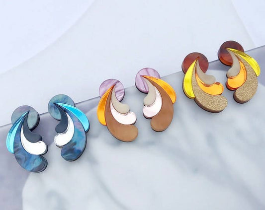 Four pairs of abstract layered stud earrings arranged on a light marble surface. The earrings feature curved, petal-like layers in different color combinations: one pair is blue, white, and gray; two pairs are orange, beige, and brown; and two pairs are yellow, orange, brown, and white.