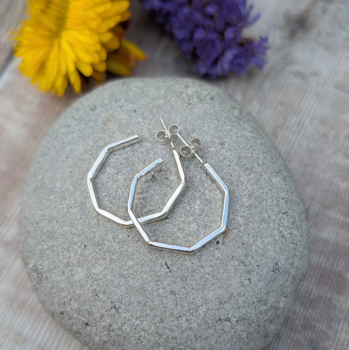 A pair of silver-toned geometric hoop earrings with openwork cutouts, displayed on a smooth gray stone with blurred yellow and purple flowers in the background.