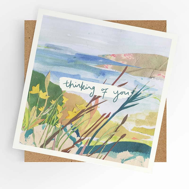 A square art print card with a white border, featuring a watercolor landscape of coastal dunes and reeds with yellow flowers and the handwritten text 'thinking of you', displayed on top of a brown kraft envelope against a white background.