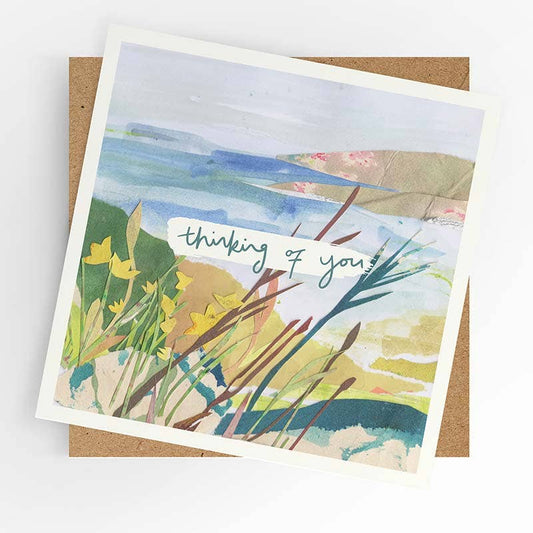 A square art print card with a white border, featuring a watercolor landscape of coastal dunes and reeds with yellow flowers and the handwritten text 'thinking of you', displayed on top of a brown kraft envelope against a white background.