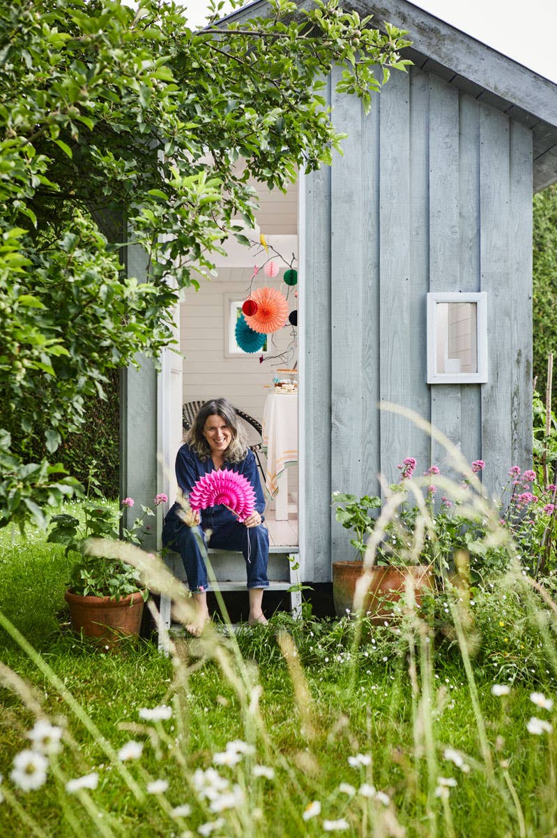 Woman sitting in a garden shed holding a pink flower, surrounded by greenery and flowers.