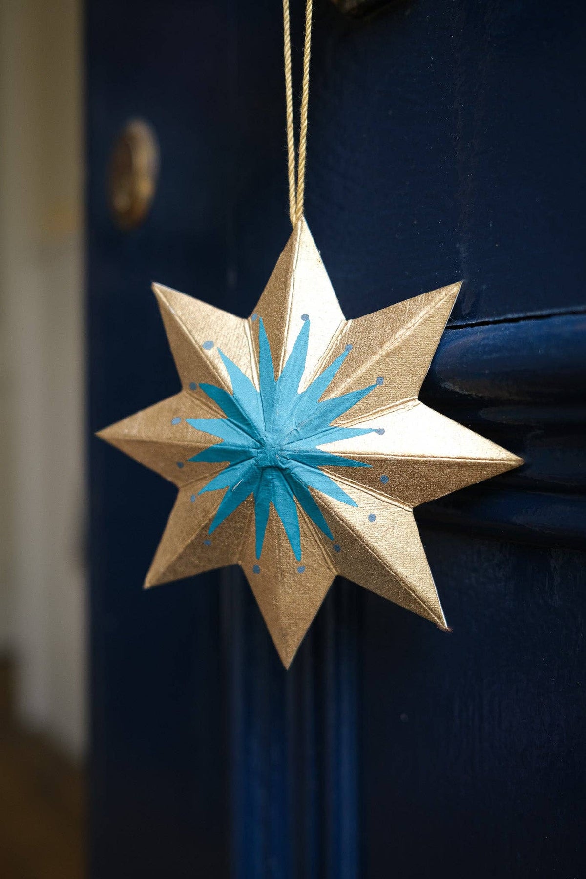 A metallic gold eight-pointed star ornament with a bright blue starburst pattern in the center, hanging by a gold string against a dark blue door.