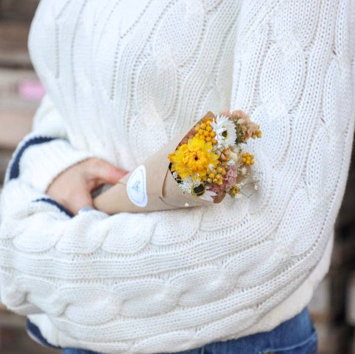 Person holding a bouquet of flowers wrapped in brown paper, wearing a white knitted sweater.