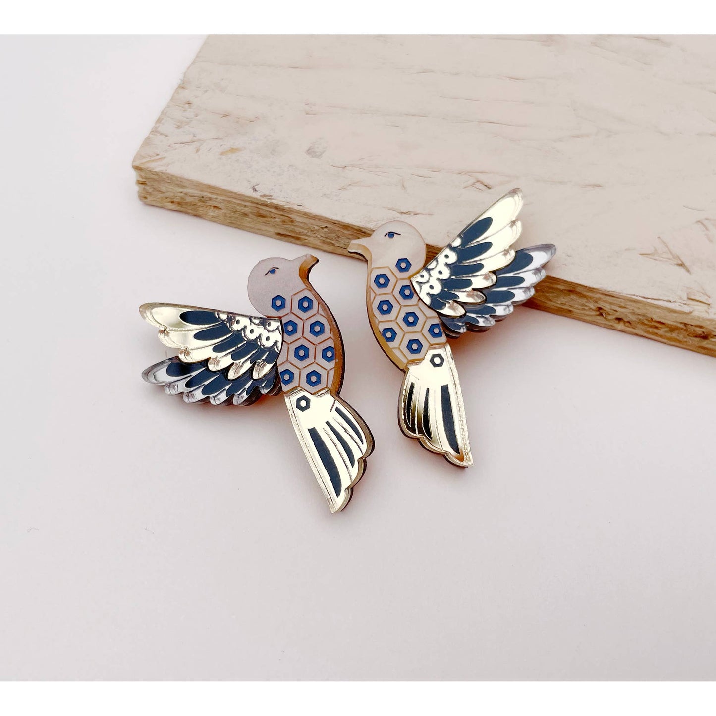 A pair of bird-shaped earrings with gold-tone metal and blue, white, and black enamel details, displayed on a light beige surface.